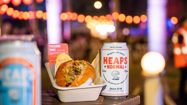 A bahn mi sits in a cardboard box next to a Heaps Normal can of beer with blurred red lights in the background