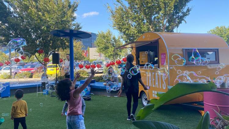 A pop-up park setting with a food truck, live musician and kids running around playing with bubbles.