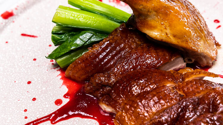 Sliced duck and a duck leg sit on a light pink plate with some greens, and red sauce splattered on the plate