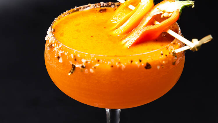 A bright orange cocktail topped with chilis on a toothpick in front of a black background