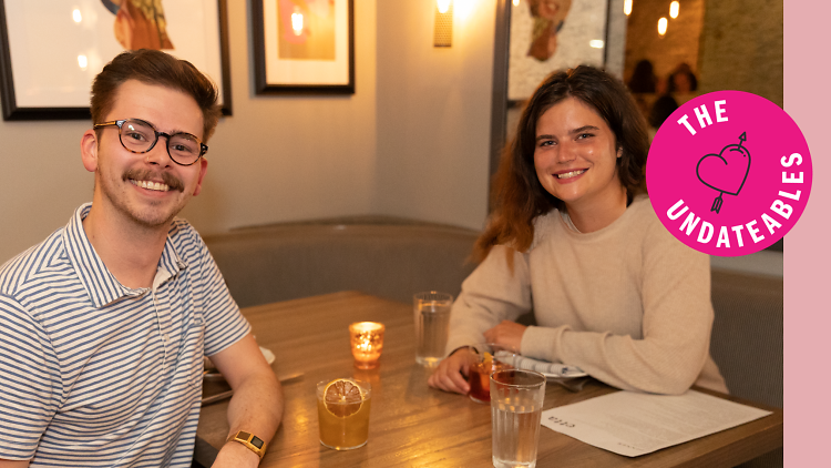 A man and a woman sit at a table with cocktails while smiling at the camera