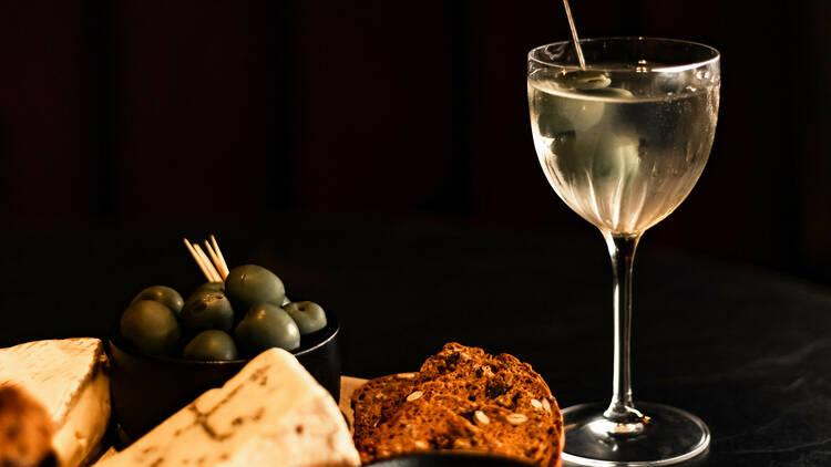 A martini sits next to a cheese plate filled with cheese crackers and olives on a black background