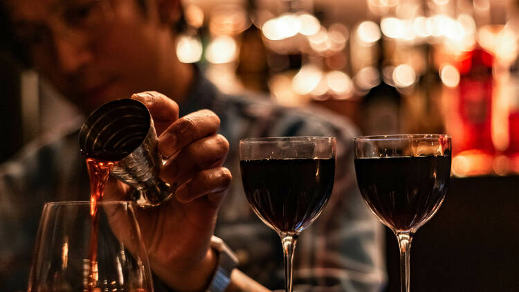 A bartender pours red liquid from a metal shaker into a short cocktail glass, which sits on the bar next to two dark coloured cocktails