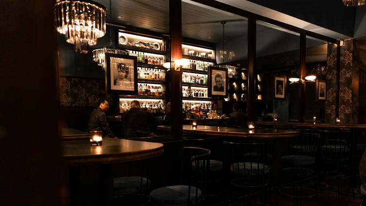 A dimly lit bar has three wooden tables against a wall covered in mirrors which reflects the backlit bar