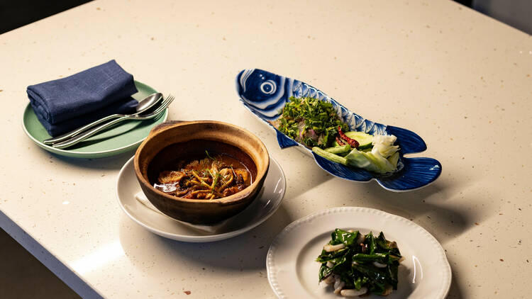 A white table with three different dishes, one of them presented in a blue dish shaped like a fish, and one in a claypot