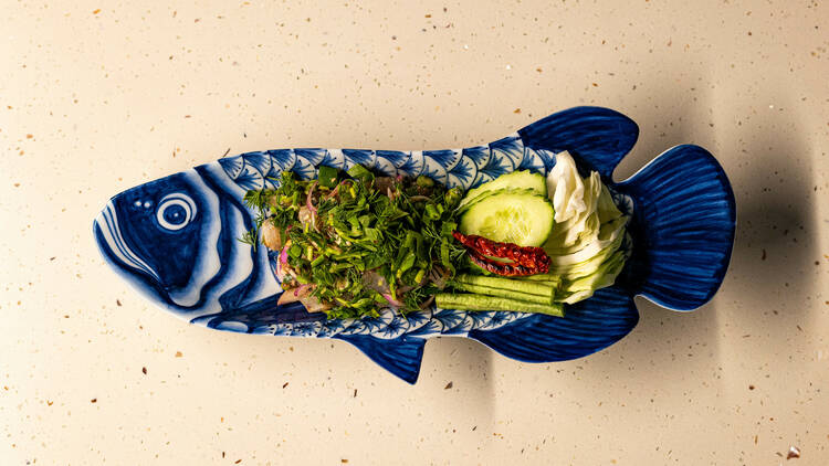 A blue fish shaped plate filled with herbs and vegetables sits on a white marble table