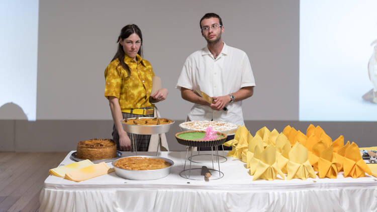 Two people stand in front of a white table covered in food