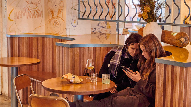 The wooden tables and benches inside Hope St Radio with two girls sitting at the top with drinks and bag of chips looking at their phone