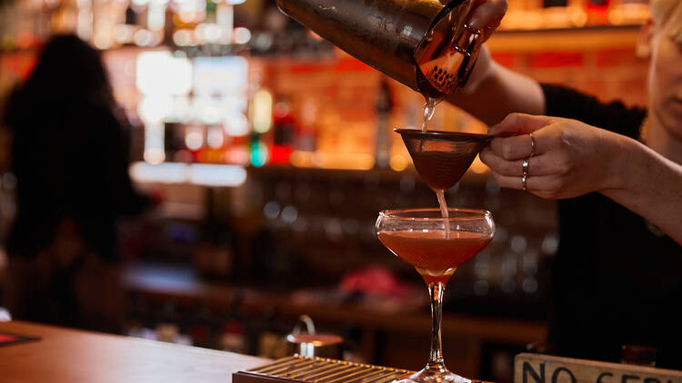A bartender pours a red cocktail from a metal shaker into a glass which sits on the bar through a strainer