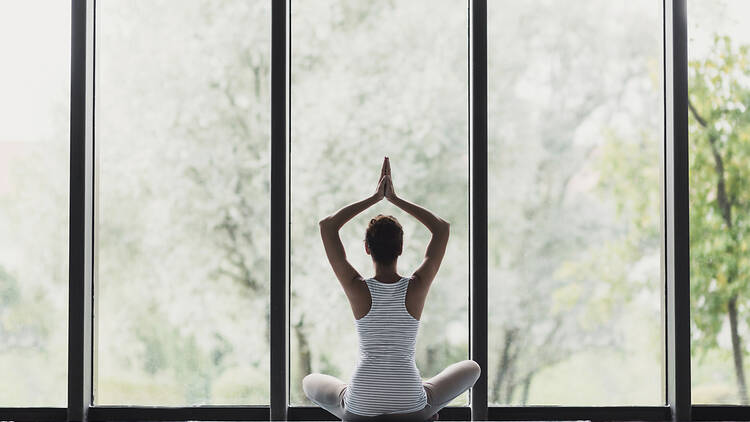 A woman in lotus pose meditating by a large glass window.