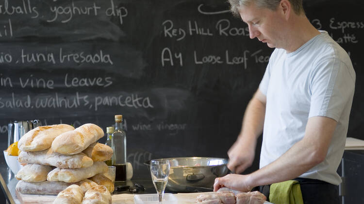 A chef cuts chicken on a chopping board next to loafs of bread and a glass of wine