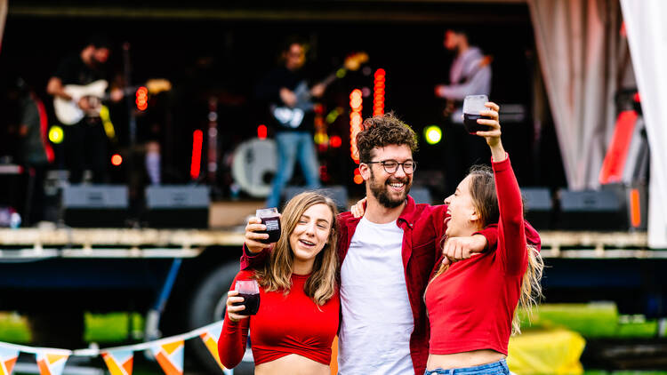 A man and two women holding glasses of red wine, enjoying an outdoor gig.