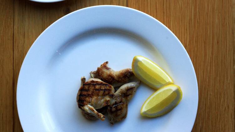 A large white plate with barbecued quail next to two lemon wedges on a wooden table