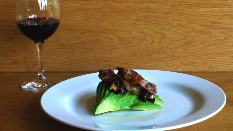 A white plate with avocado and pancetta next to a glass of red wine in front of a wooden wall