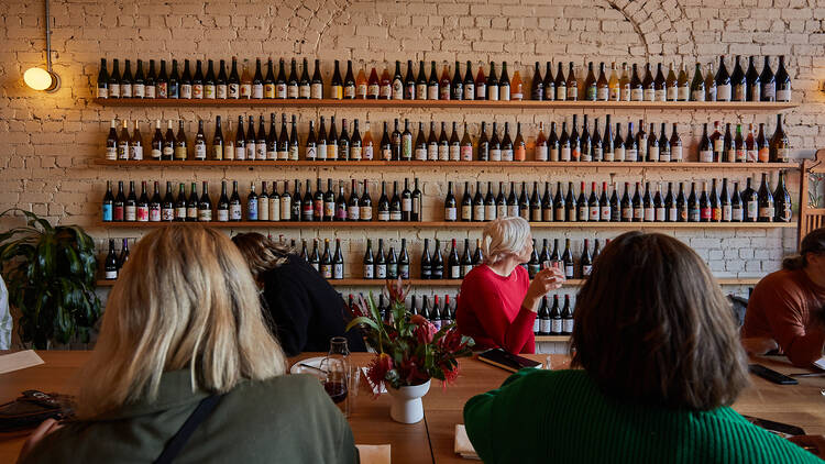 A white brick wall is filled with shelfs of wine bottles with a large wooden table with people sitting around in front of the wall