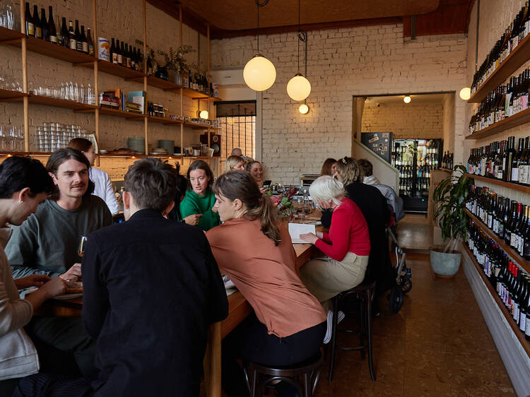 A large table in the middle of the bar is filled with groups of people eating and drinking with low hung light fittings above the table