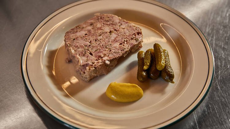 A plate with a slice of terrine, mustard and small pickled cucumbers is on a silver bench