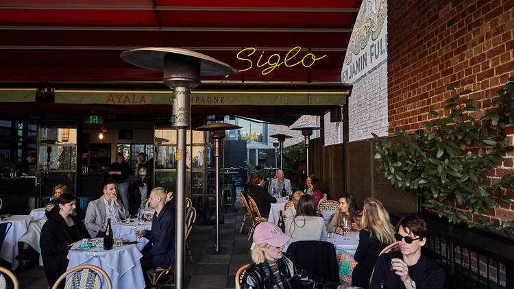 There are many groups of people sitting around small tables with white tableclothes on the deck with a small yellow sign reading Siglo above them