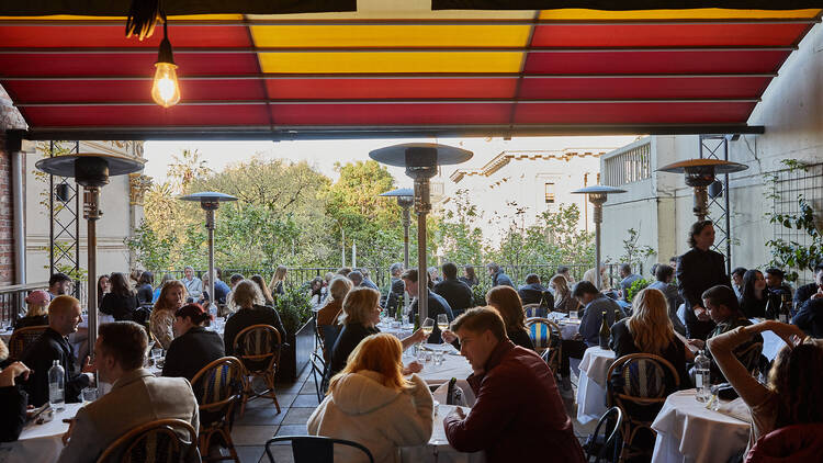There are many tables and large heaters on the deck under a red and yellow awning 