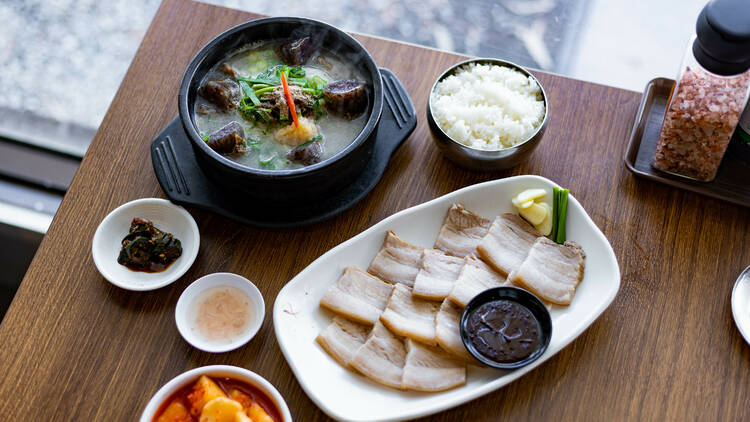 A wooden table with a hot pot, rice, sliced pork belly and small vegetables dishes