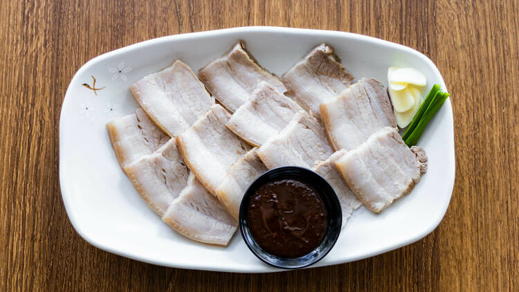 A wooden table with a white plate on top which has sliced pork belly and a small bowl of sauce