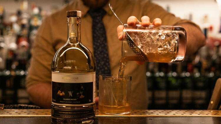 A bartender pours a cocktail from a glass mixer into a glass with a large ice cube which sits next to a bottle of whiskey on the bar