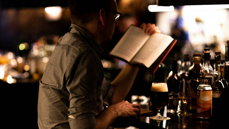 A man reads a novel and drinks a glass of beer sitting at the bar