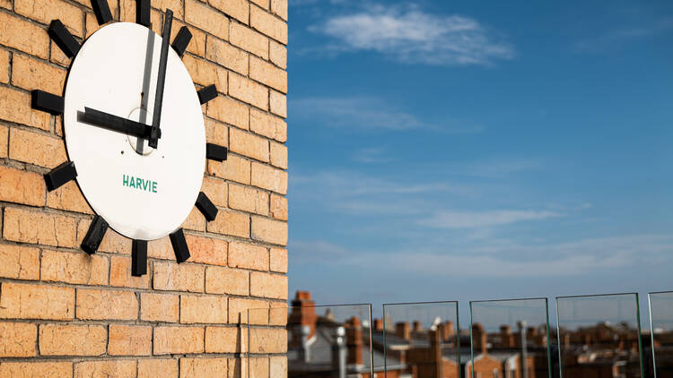 A large white clock on a brick wall on the rooftop bar with blue skies in the background