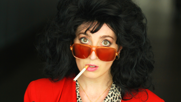 A woman in a black puffy wig and red lense sunglasses has a cigarrete hanging out her mouth