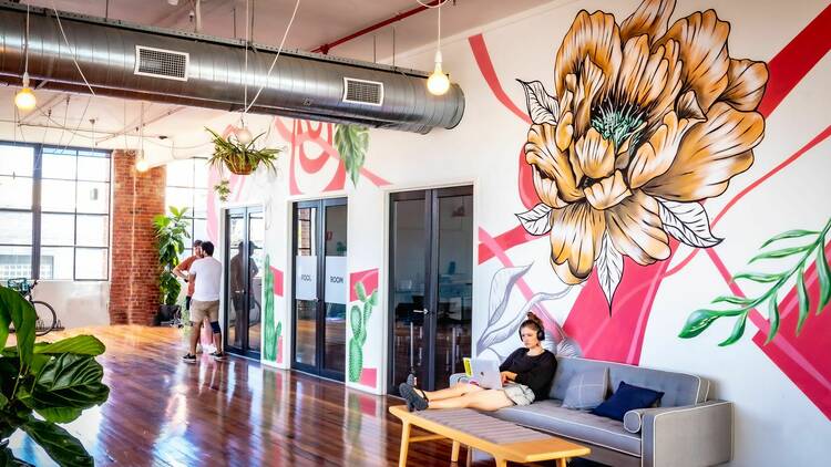 A woman sitting on a couch against a giant mural of a peony, in a co-working space.