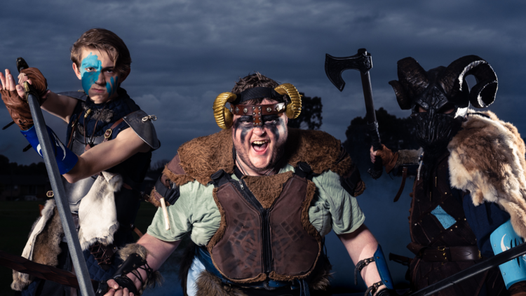 Three people holding weapons in fancy medieval dress action pose against a stormy sky