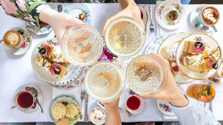 An overhead shot of four people cheersing with Champagne above a table laden with high tea goodies.