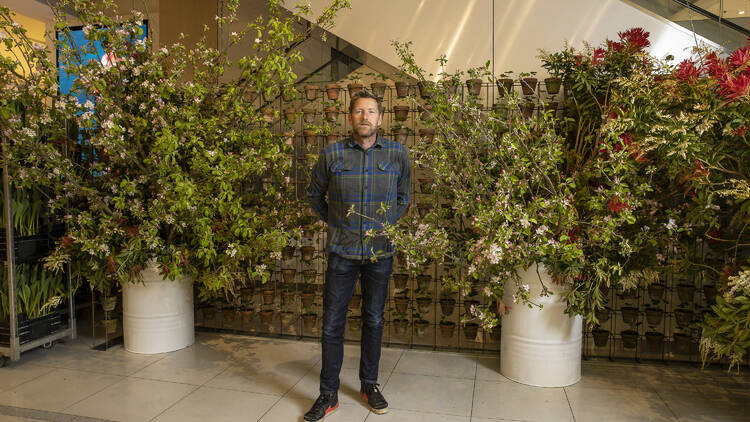 A man in a check shirt stands in front of a large, leafy installation.