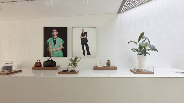 The indoor store has a white bench with different accessories spread out and a few plants with two framed pictures on the wall 