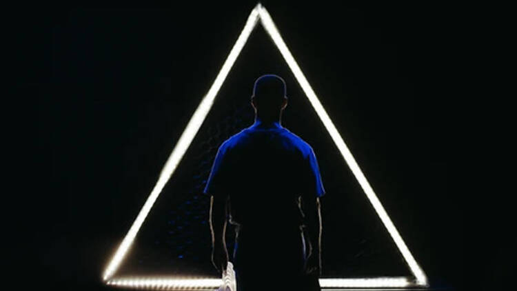 A dark room with a male figure facing an illuminates neon triangle.