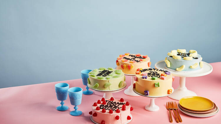 A pink table in front of a blue wall has 4 different colourful cakes on it with blue cups and plates