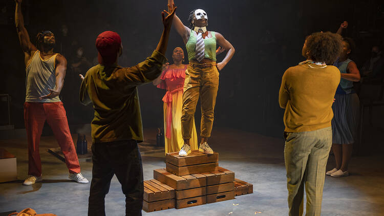 A person in a white mask stands on a podium with their arm raised surrounded by other people with their arms raised