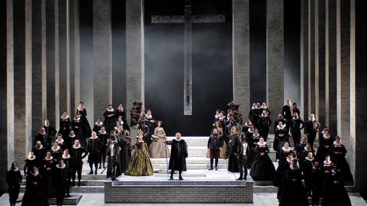 Performers in black outfits stand on a large stage during an opera performance of Don Carlos