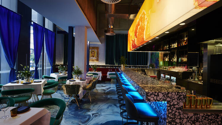 Inside Bombay Yacht Club there are bright blue floors and curtains, an orange mural above a long bar with blue stools,