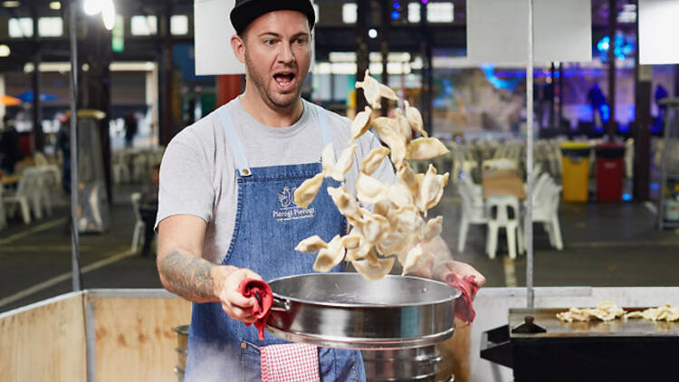 A chef in an apron and a black cap tosses a bowl of pierogi dumplings into the air