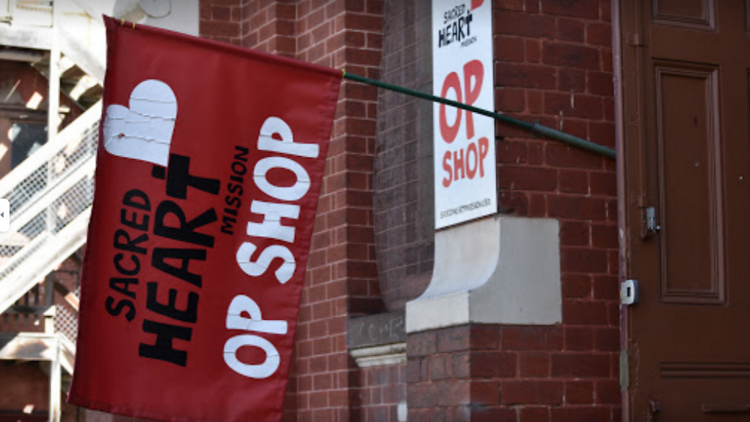 A red flag hoisted from a brick building, reading 'Sacred Heart Mission Op Shop'.