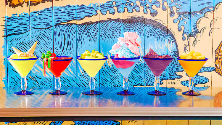 A line of six different margarita flavours topped with lollies and fairy floss in front of a bright mural painting on the wall 