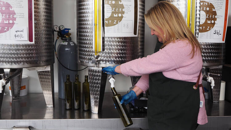 A lady in an apron and pink jumper pours olive oil out of a large vat into a bottle