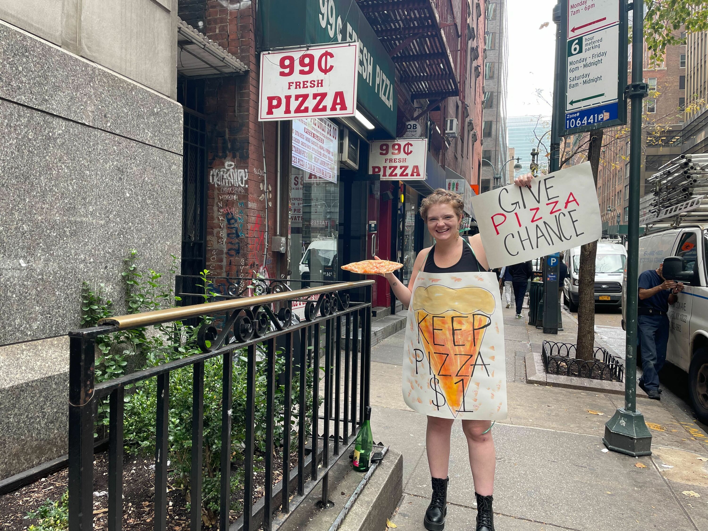 A woman wears a sandwich board reading "Keep Pizza $1." 