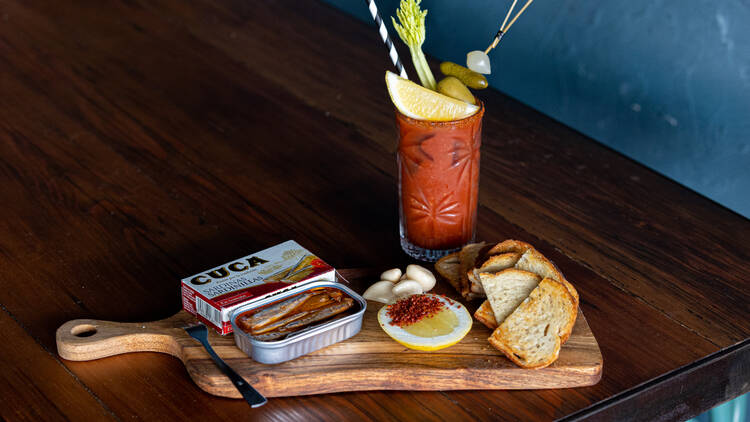 A wooden table with a blood mary on it, next to a wooden board with a tin of anchovies and toasted bread