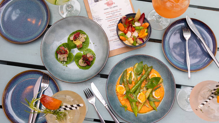 A table has blue plates, cocktails, and three colourful plates of food from a birds eye view