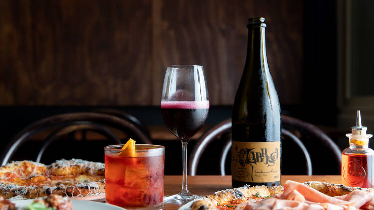 A table has two pizzas, a negroni, a bottle of wine and a glass of red wine on it