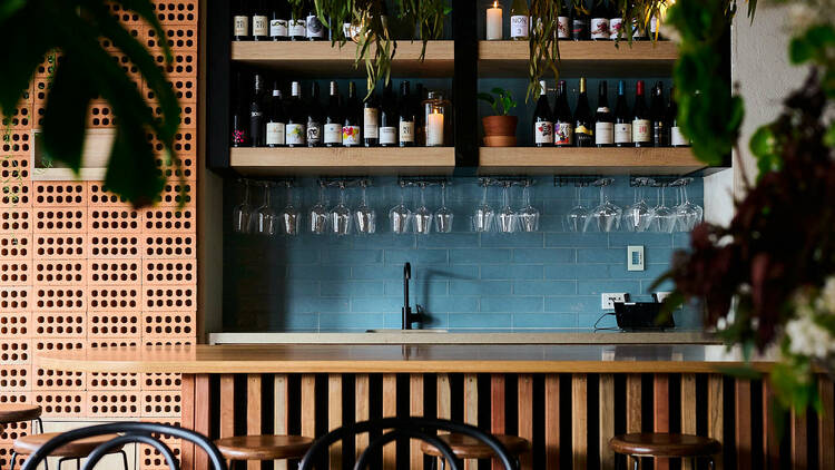 The bar at Nazar has wooden bar top, wooden stools, hanging plants and shelves of wine in front of blue tiles