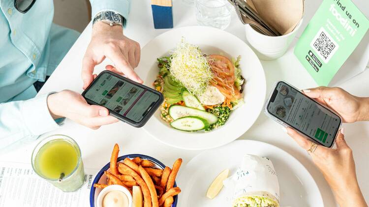 Two people at a table have their phones out using the Foodini app with three plates of food on the table including fries, a wrap and a salad