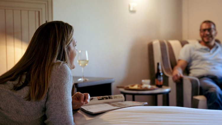 A woman holding a wine glass smiles on a bed at a man in a chair 